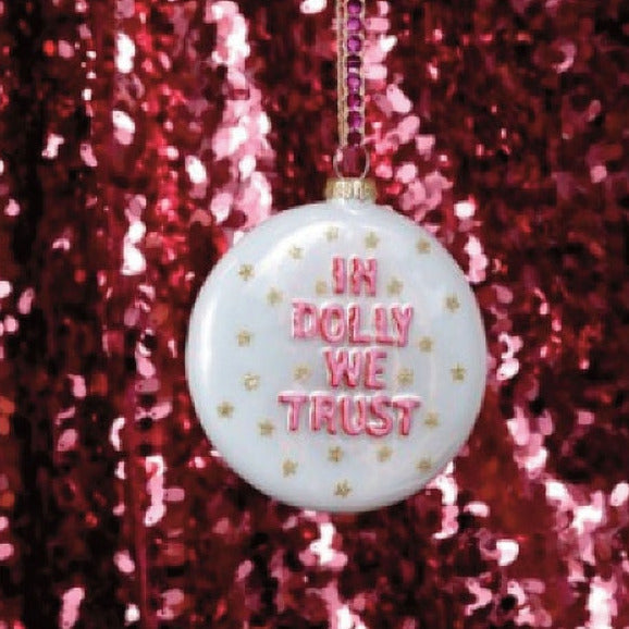 A disk-shaped white glass Christmas ornament with pink text reading 'IN DOLLY WE TRUST' and small gold stars, hanging from a tree branch against a sparkling red sequin backdrop.