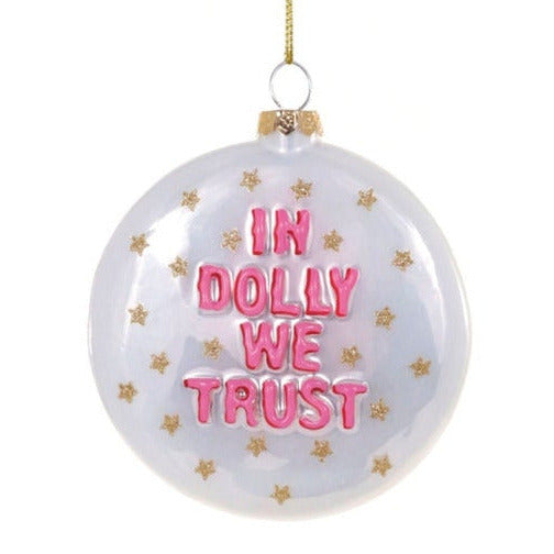 A disk-shaped white Christmas ornament with pink raised text reading 'IN DOLLY WE TRUST' and small gold star accents, hanging from a gold string against a white background.