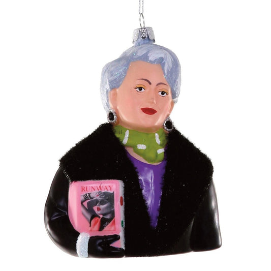 Decorative ornament pictures a glamorous woman holding a copy of Runway magazine. She has cropped silver hair and wears a black leather coat with faux fur collar over a purple dress. Her large earrings, green scarf, and glitter-cuffed black gloves complete the look.