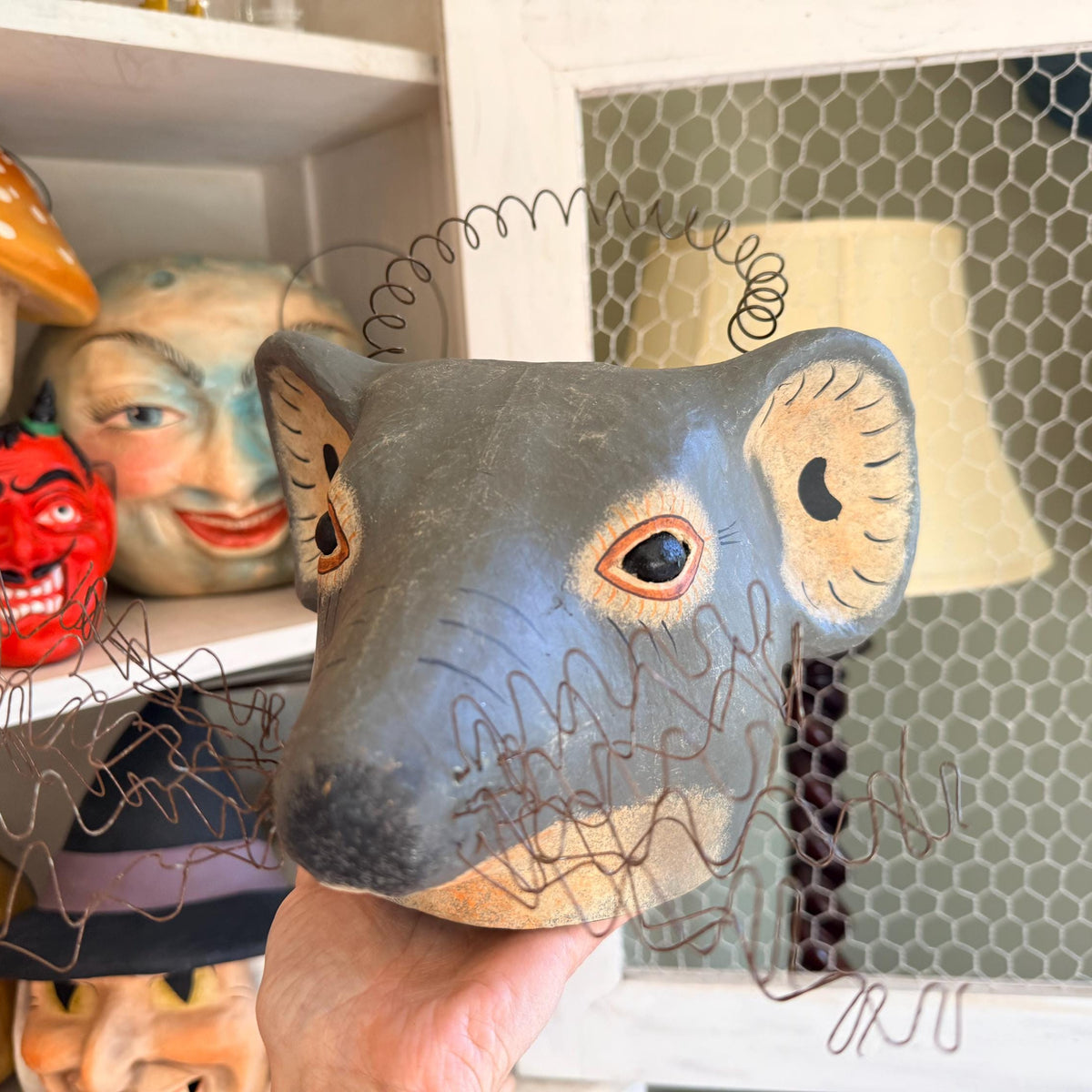 A hand holds a hand-painted paper mache mouse bucket in front of a shelf with other colorful folk art buckets and a window with wire mesh.
