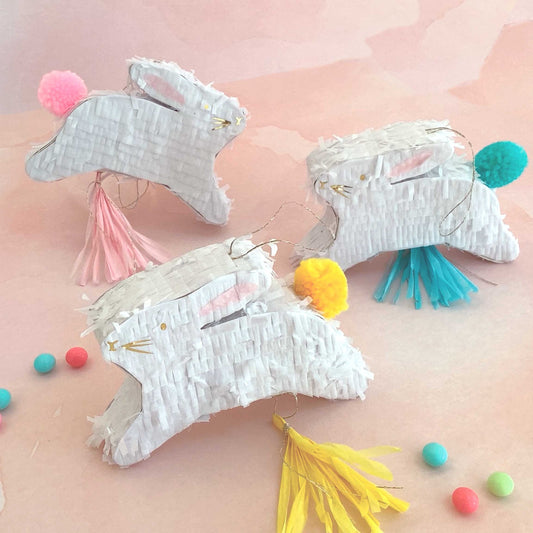 Three white-fringed boxes shaped like bunnies with pom pom tails and tassels in pink, yellow, and blue on a pink background.