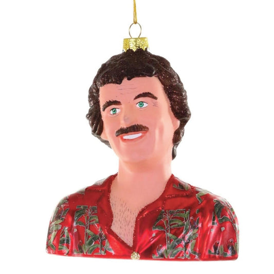 A novelty Christmas ornament shaped like a bust of a hunky man with dark curly hair, a mustache, green eyes, and a hairy chest, wearing a red Hawaiian shirt with a green tropical pattern. The ornament hangs from a gold string against a plain white background.