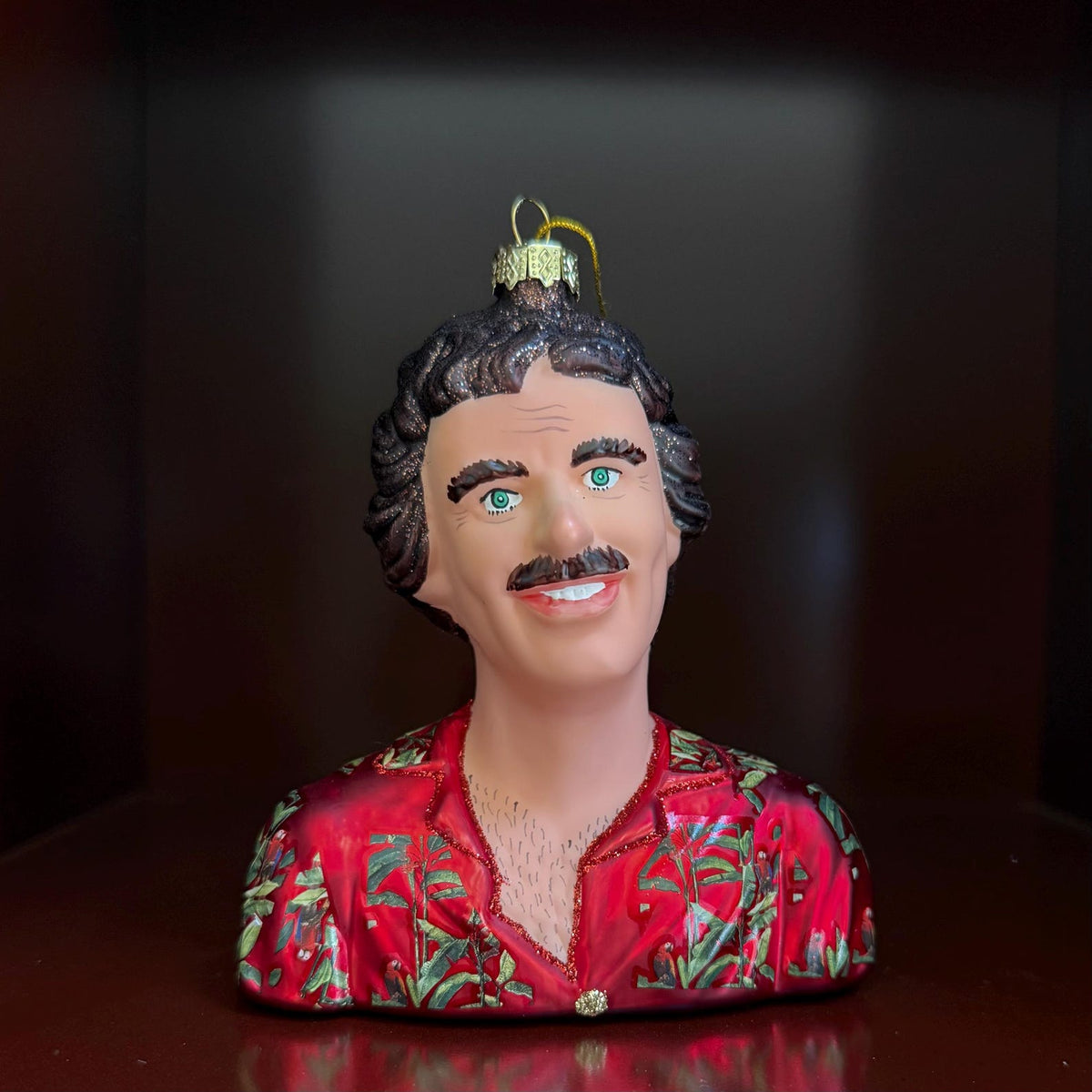 A novelty bust-shaped ornament or figurine of a smiling man with dark hair, bushy eyebrows and mustache, green eyes, and a red tropical-patterned shirt opened to reveal a hairy chest, shown against a dark background.