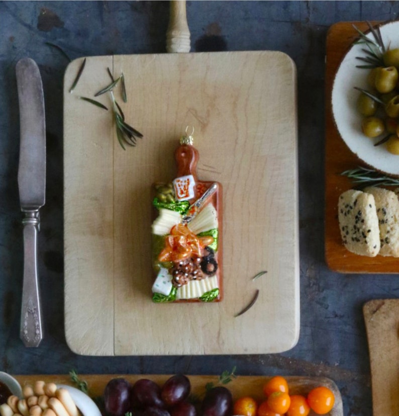 Wooden cutting board with a decorative charcuterie board ornament filled with cheese and various food items on a table surrounded with a knife and other buffewt trays.