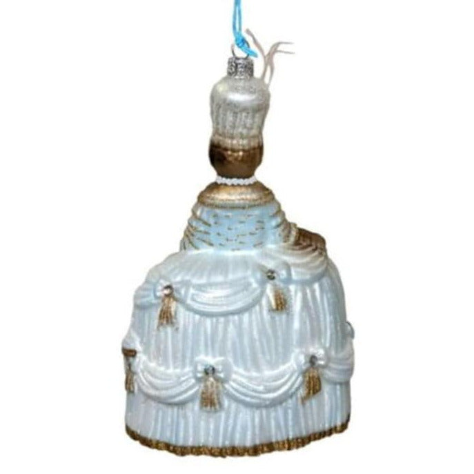 A vintage-style Christmas ornament shaped like a woman wearing a fancy draped dress, with a white and light blue body, gold trim, tassels, and a blue hanging string on a plain white background.
