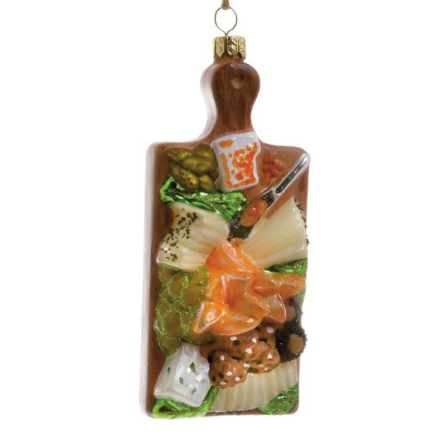 Decorative ornament shaped like a wooden cutting board with cheese, pickles, and various food items on a white background