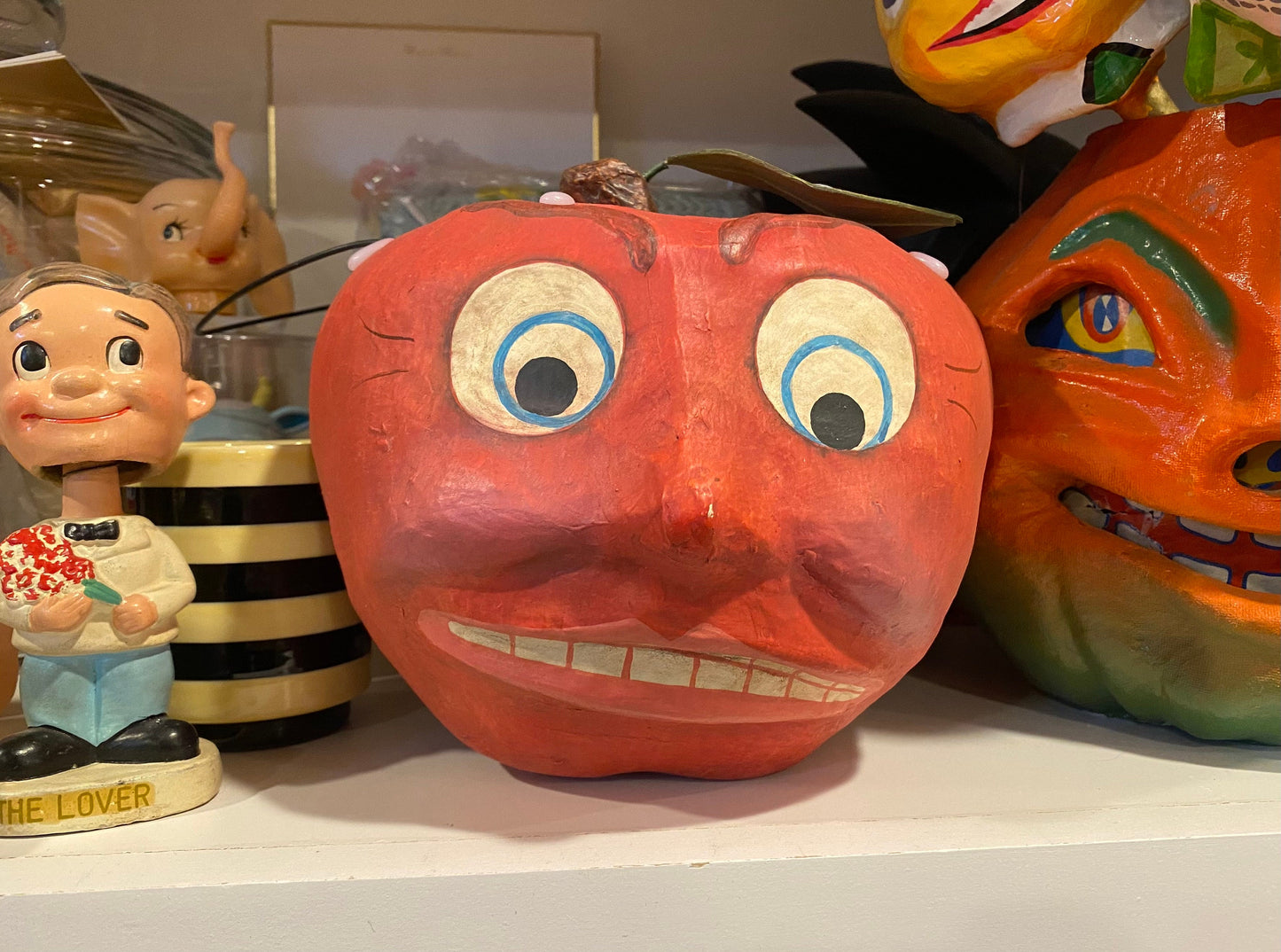 An orange hand-painted wooden pumpkin with a whimsical face, featuring large blue-rimmed eyes and a toothy grin, displayed on a shelf alongside other decorative figurines.
