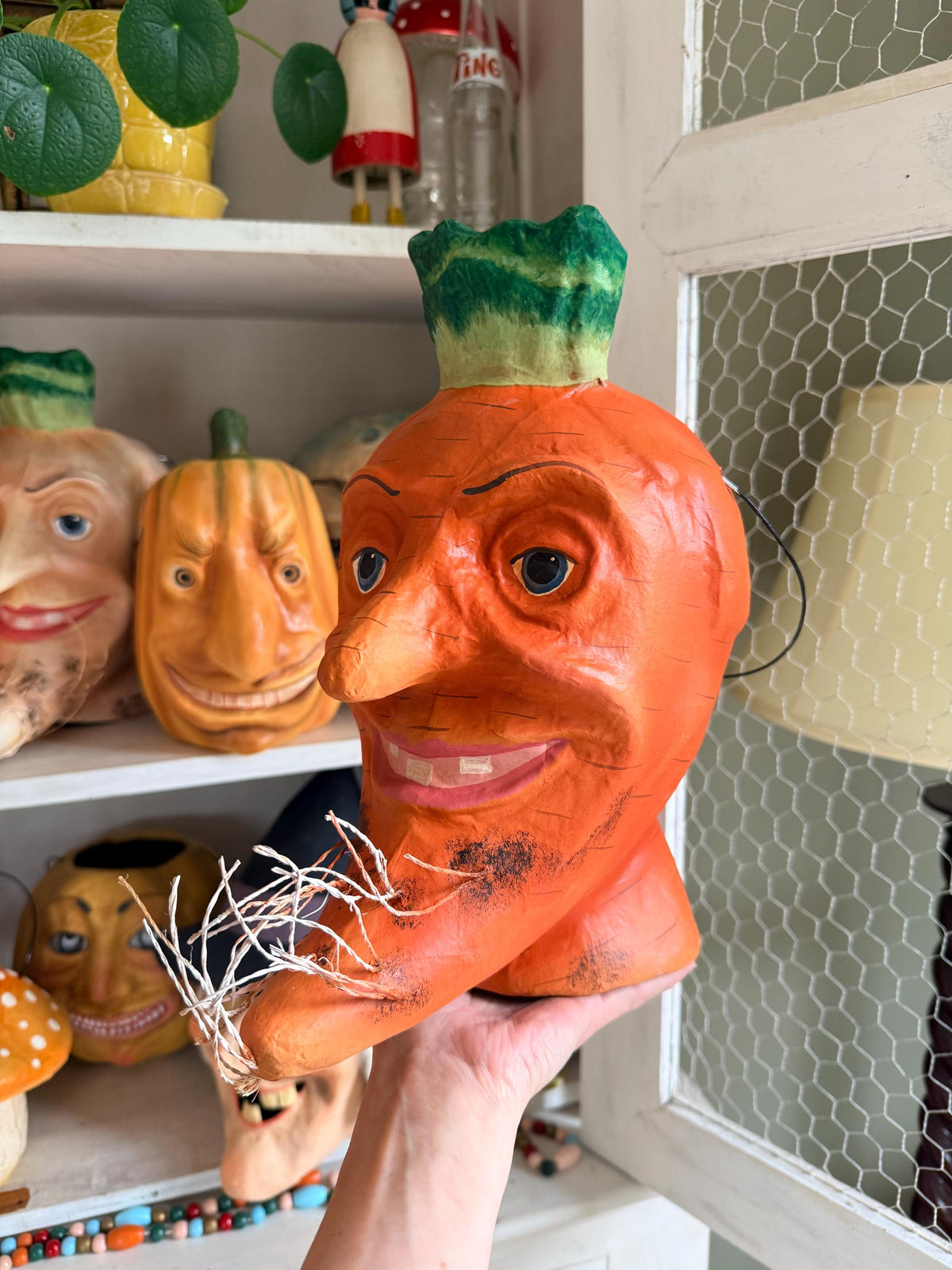 A hand holds a large, novelty carrot-shaped head decoration with a painted orange body, green leafy top, and a straw -dotted chin in a cabinet with shelves of other decorative items.