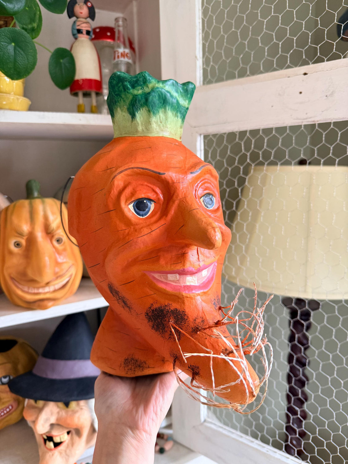 A hand holds a carved orange carrot head bucket with a green leafy top and blue eyes, photographed indoors on a shelf next to a lamp and other decorative items.
