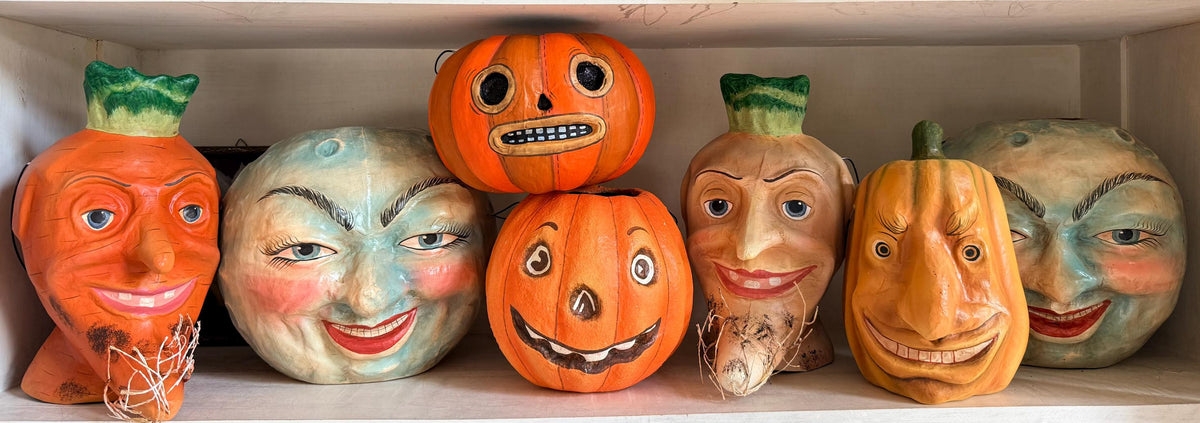 A collection of hand-painted paper mache Halloween buckets displayed on white shelves, including three jack-o'-lanterns with human faces, a carrot-shaped head, and a green mushroom cap.