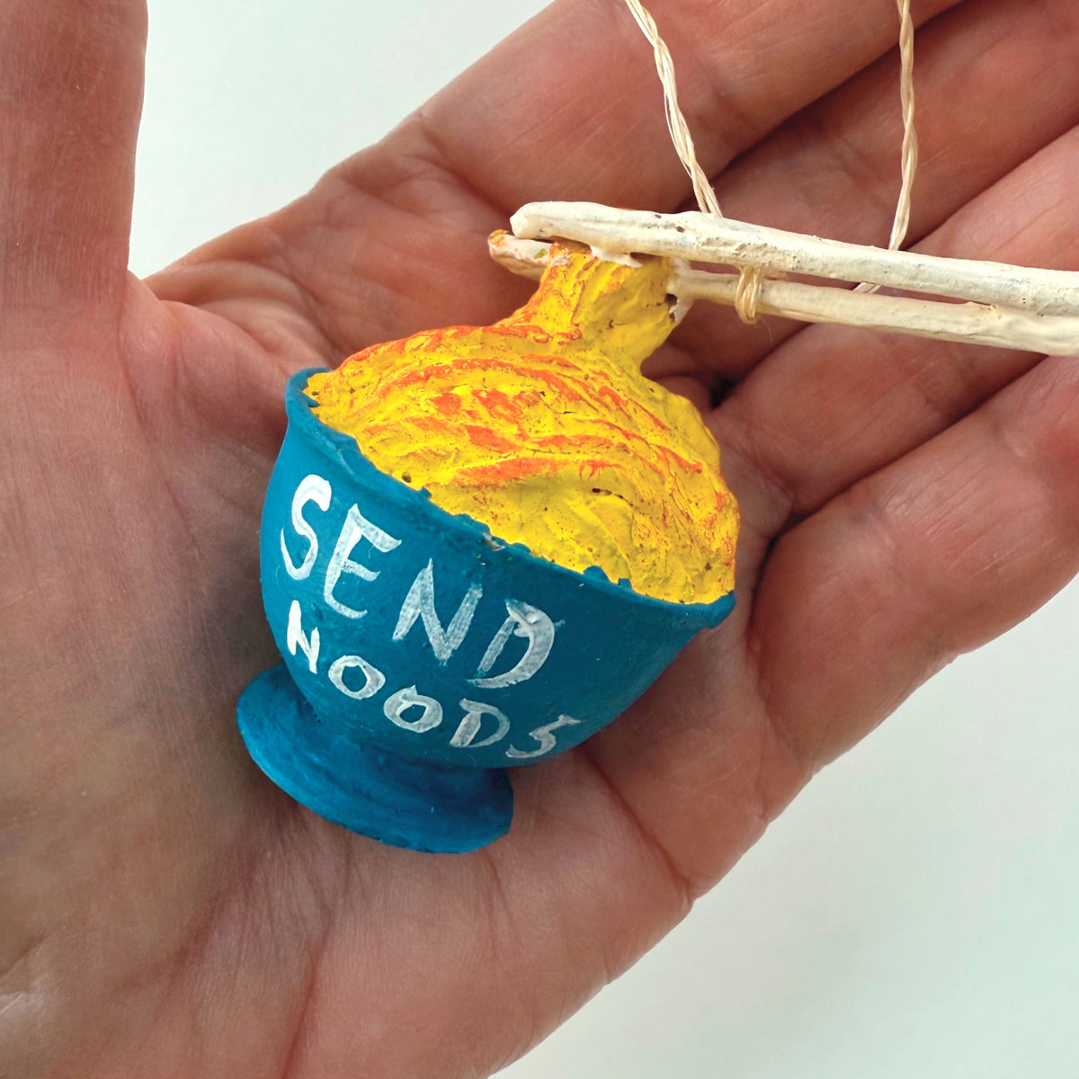 A miniature ceramic ornament shaped like a bowl of noodles with chopsticks resting on top, held in a person's hand. The bowl is bright blue with the words 'SEND NOODS' in white, filled with yellow and orange painted noodles, and hangs from a thin string.