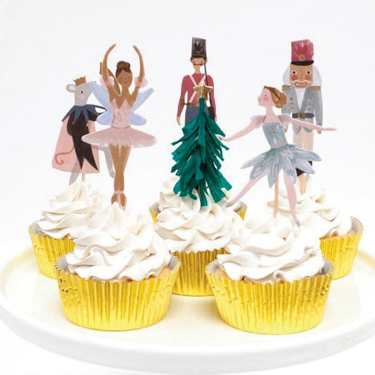 Six cupcakes topped with picks representing characters from the Nutcracker Ballet as well as a tissue fringed Christmas tree.  Cupcakes are in gold foil liners and picks are placed atop fluffy while frosting.