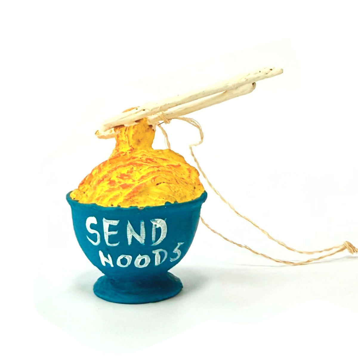 A novelty yornament designed to look like a bowl of noodles, with a blue bowl base, yellow textured pasta strands, wooden chopsticks on top, and the words 'SEND NOODS' printed in white on the side. It is shown on a clean white background.